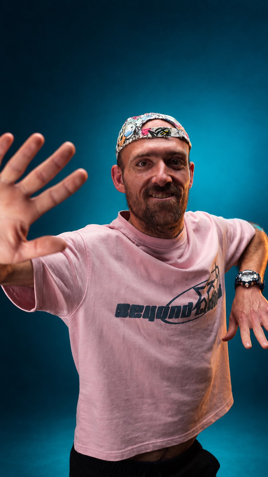 Marius Els in 'n pienk t-hemp en bandana, arm uitgestrek na die kamera, teen 'n diep petrol-blou agtergrond